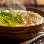 Imagine a close-up shot of a steaming bowl of savory oatmeal. The oatmeal is topped with slices of perfectly ripe avocado, sprinkled with crunchy toasted sesame seeds, and drizzled with a swirl of fragrant sesame oil. The bowl rests on a rustic wooden tabletop, with a vintage spoon on the side. The lighting is soft, creating a cozy and inviting atmosphere.